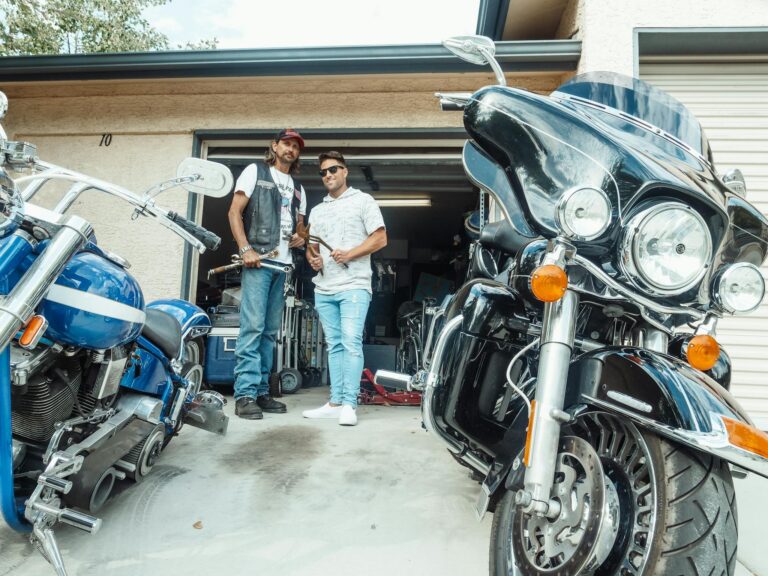 Two men standing with tools in a garage workshop surrounded by motorcycles.